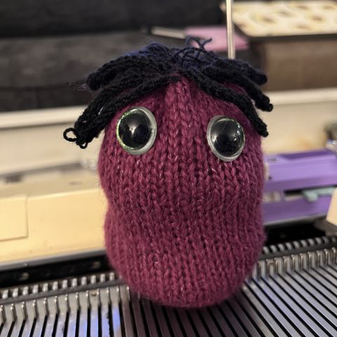 A stuffed lil guy made of red fuzzy yarn, with big green plastic eyes and thick hair made of black yarn. It's sitting on a knitting machine bed.