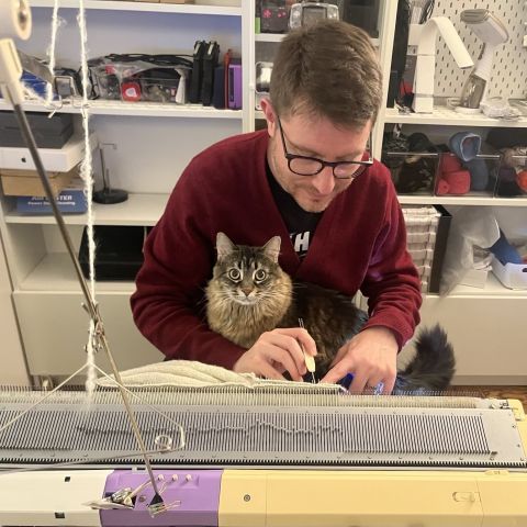 Lolly the cat makes eye contact with the camera as she sits in Marty's lap. Marty is focused on hanging stitches on his knitting machine.