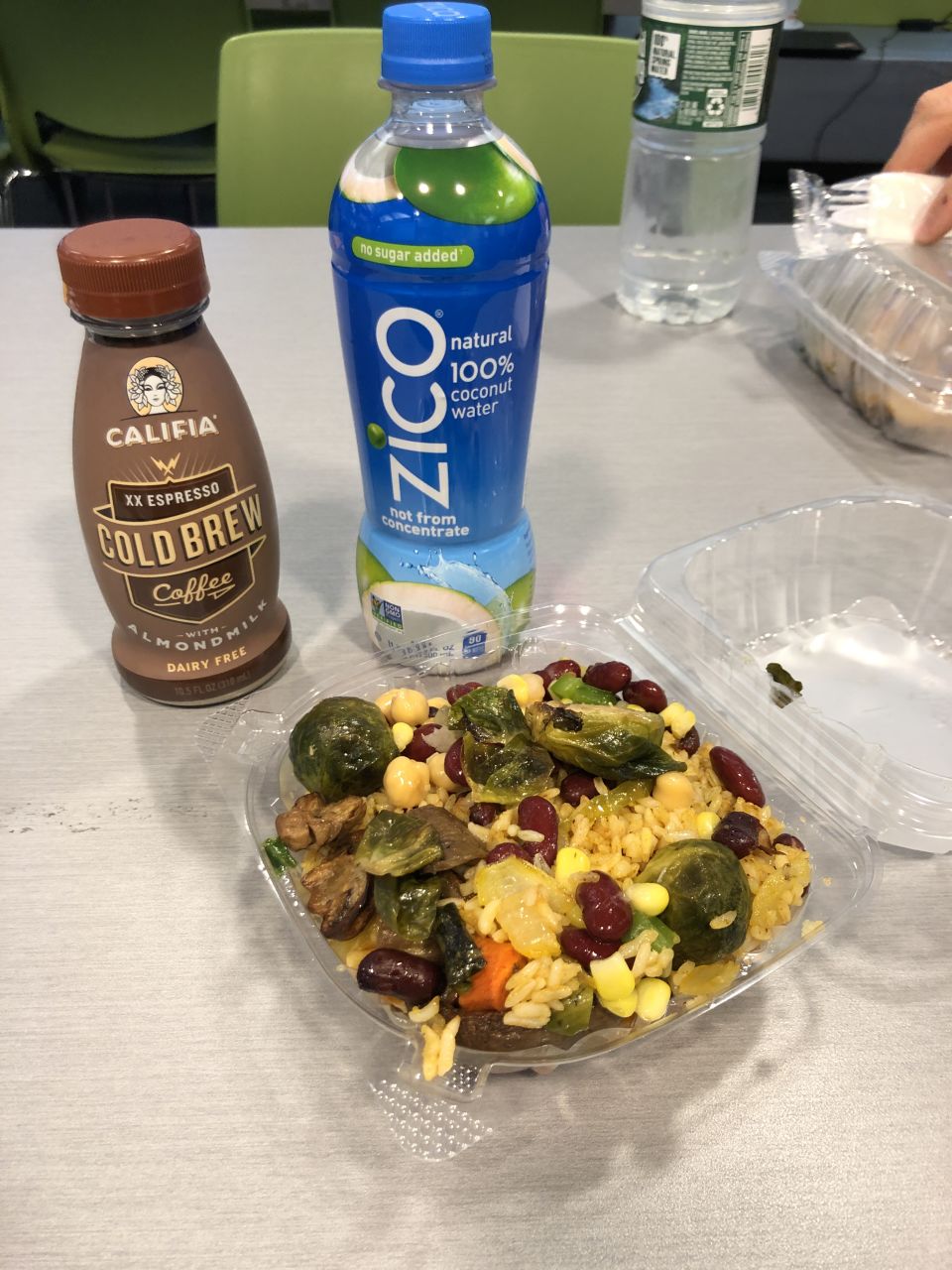 a bottle of cold brew coffee, a bottle of coconut water, and a plastic clamshell full of rice, beans, and brussels sprouts