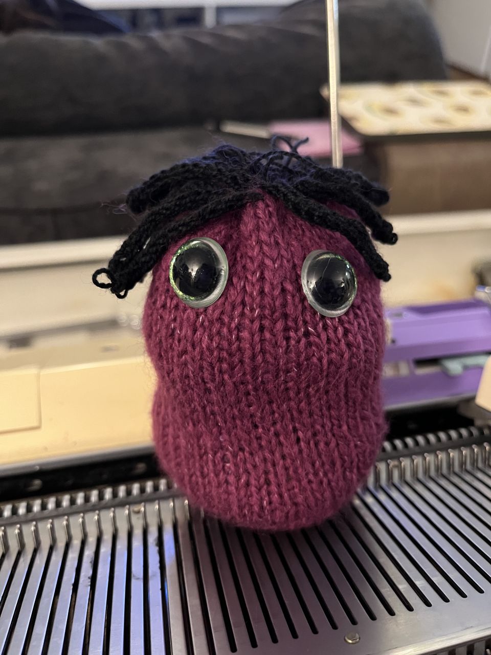 A stuffed lil guy made of red fuzzy yarn, with big green plastic eyes and thick hair made of black yarn. It's sitting on a knitting machine bed.