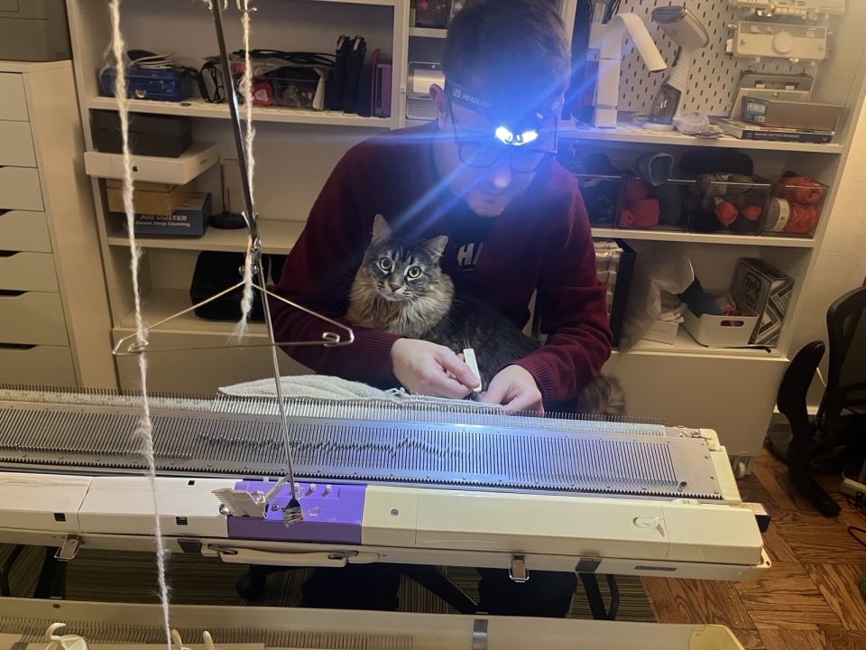 Lolly the cat sits in Marty's lap as he works stitches on his knitting machine. She is dramatically lit by the headlamp on his head.