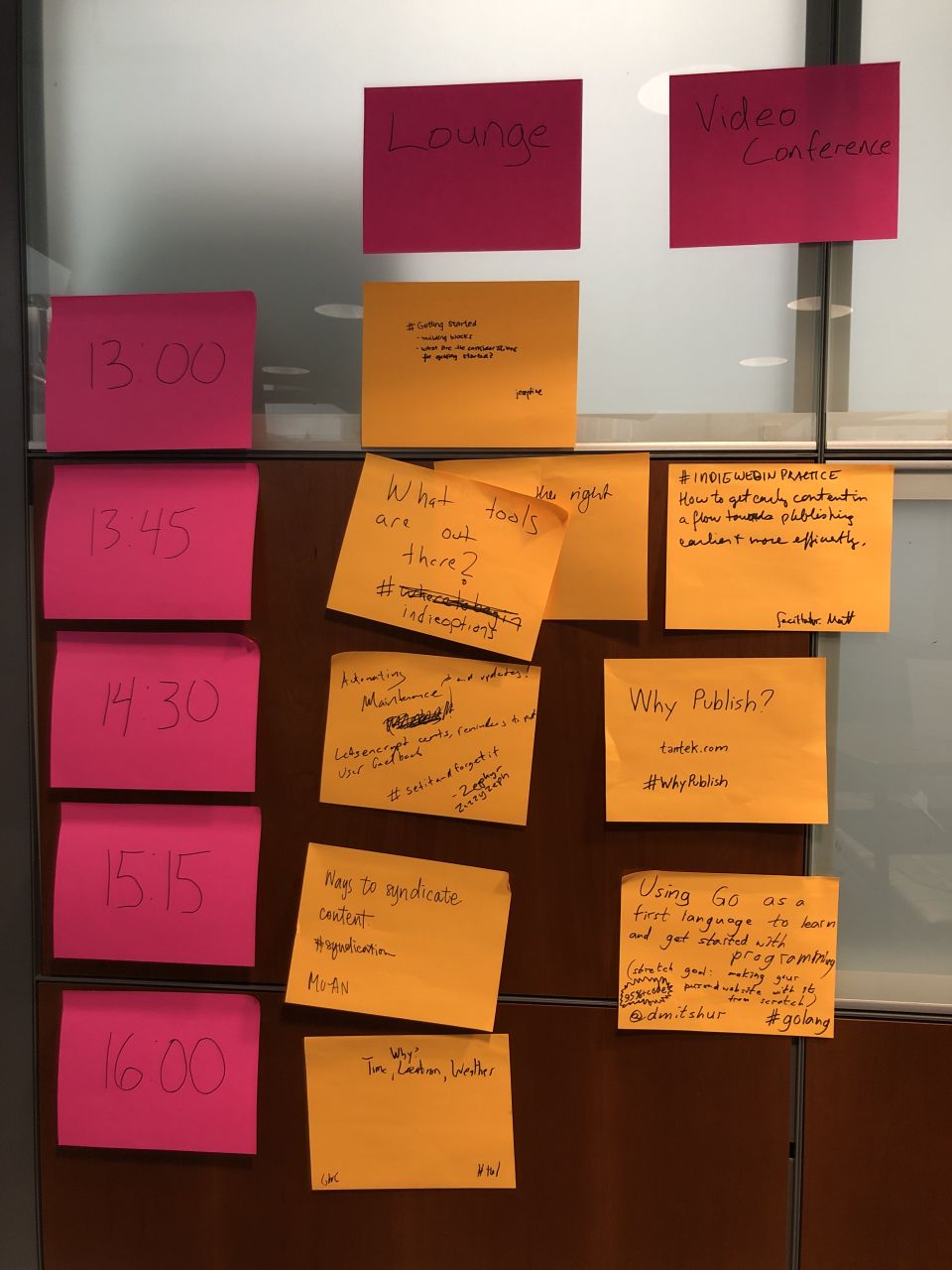 wood and glass cubicle wall covered in pink and orange sticky notes indicating times, rooms, and topics to discuss