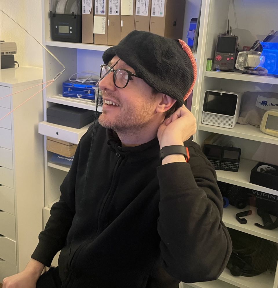 me posing awkwardly while holding the partially finished Chattie around my head. Sideways stitches are visible in the black yarn that the hat is made of, as well as sections of pink stitches in waste yarn at the edges.