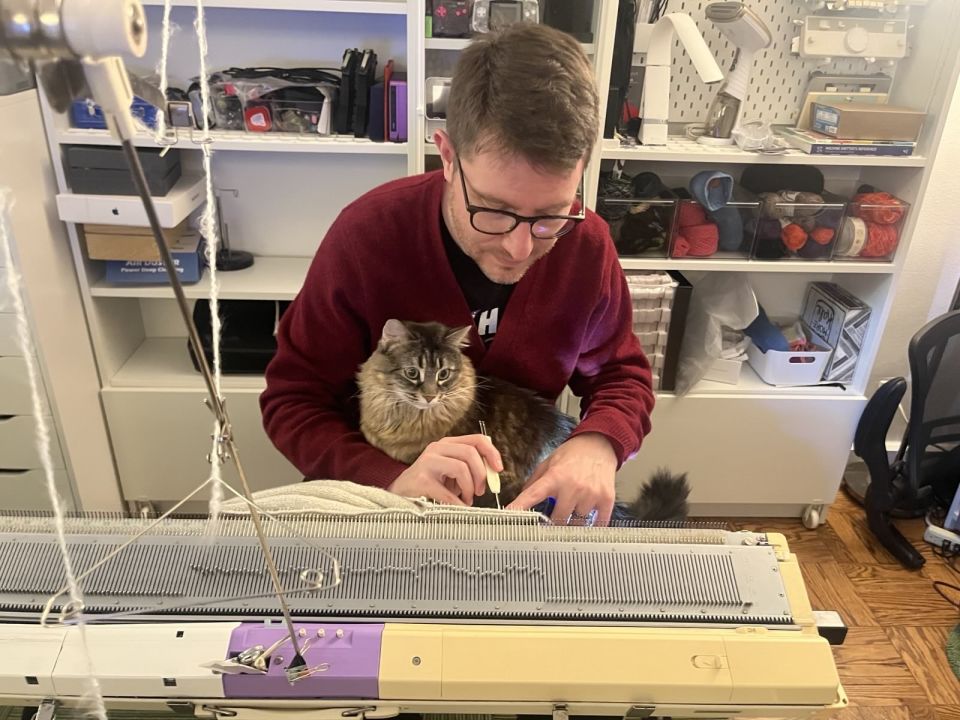 Marty works stitches on his knitting machine around Lolly the cat, who is sitting in his lap and watching him work.