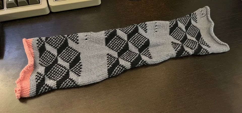 Knit swatch with three patterned sections in gray and black yarn. Each section uses the tiling cube pattern and is separated by gray areas with lace holes that indicate the tension used to knit that section.