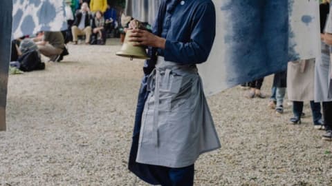 A man holds a bell standing on rocky gravel
