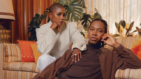A portrait-style photograph of two people in a living-room type area. One is sitting on a couch; the other is sitting in front of the couch.
