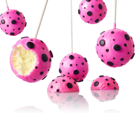 Closeup color photograph of cake pops suspended in a white space, reflecting in a white surface below. They are dipped in pink frosting with chocolate dots. One of the cake pops has been bitten into, revealing the spongy interior.
