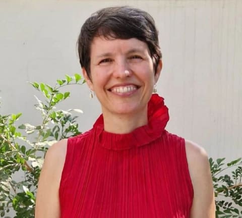 A headshot of a woman in a red blouse in front of a beige wall and green plants (Nirith Nelson)