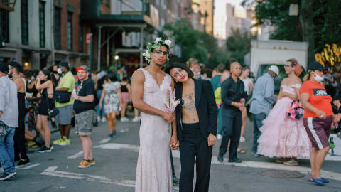 Two people are holding each other in the middle of a crowded street. One person is wearing a dress with a flower crown and the other a suit.
