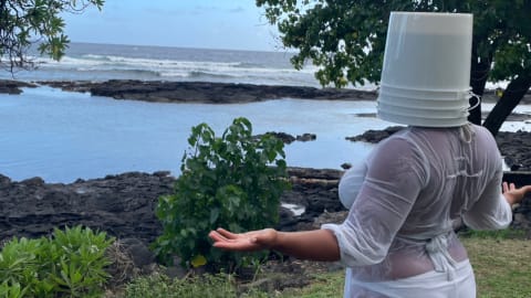 person wearing white standing next to a body of water with a white bucket on their head and arms bent out to sides.