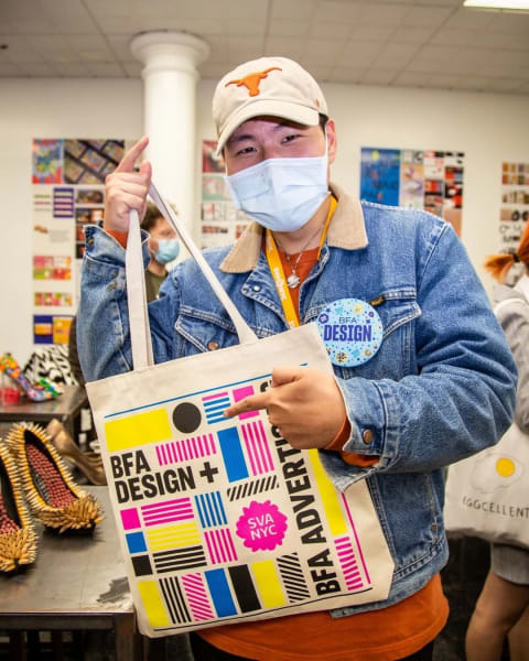 A young man stands with a BFA Design tote bag that he's holding up and pointing to. He is wearing a jean jacket and a baseball cap