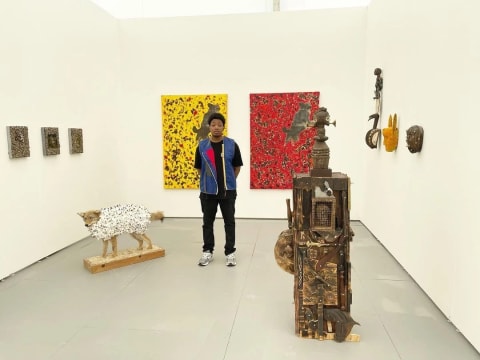 A young man stands in a white-wall gallery room filled with sculptures and paintings. Next to him is a taxidermy coyote with cotton buds covering it's body, behind him are two large paintings, one red and the other yellow