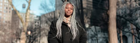 A young Black student with long grey braids wearing a black pea coat. The background shows city buildings and trees with no leaves on them.