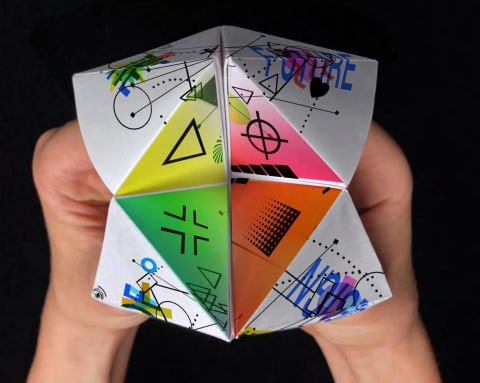 A photograph of hands holding a white paper chatterbox, aka, a "paper fortune teller."