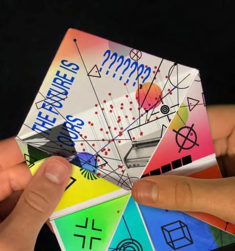 A photograph of hands holding a white paper chatterbox, aka, a "paper fortune teller."