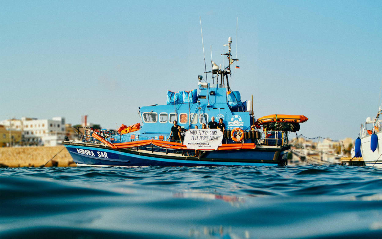 Ein Schiff von Sea-Watch im Mittelmeer. Vorne sind einige Wellen zu sehen, und im Hintergrund einige Häuser, da das Boot sich in Küstennähe befindet.