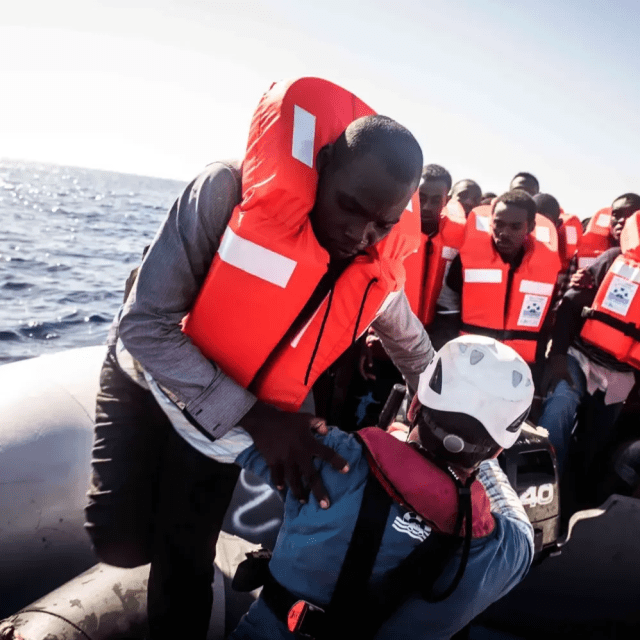 A Sea-Watch crew member wearing a helmet helps a rescued person wearing a life jacket on an overcrowded rubber dinghy in the Mediterranean.