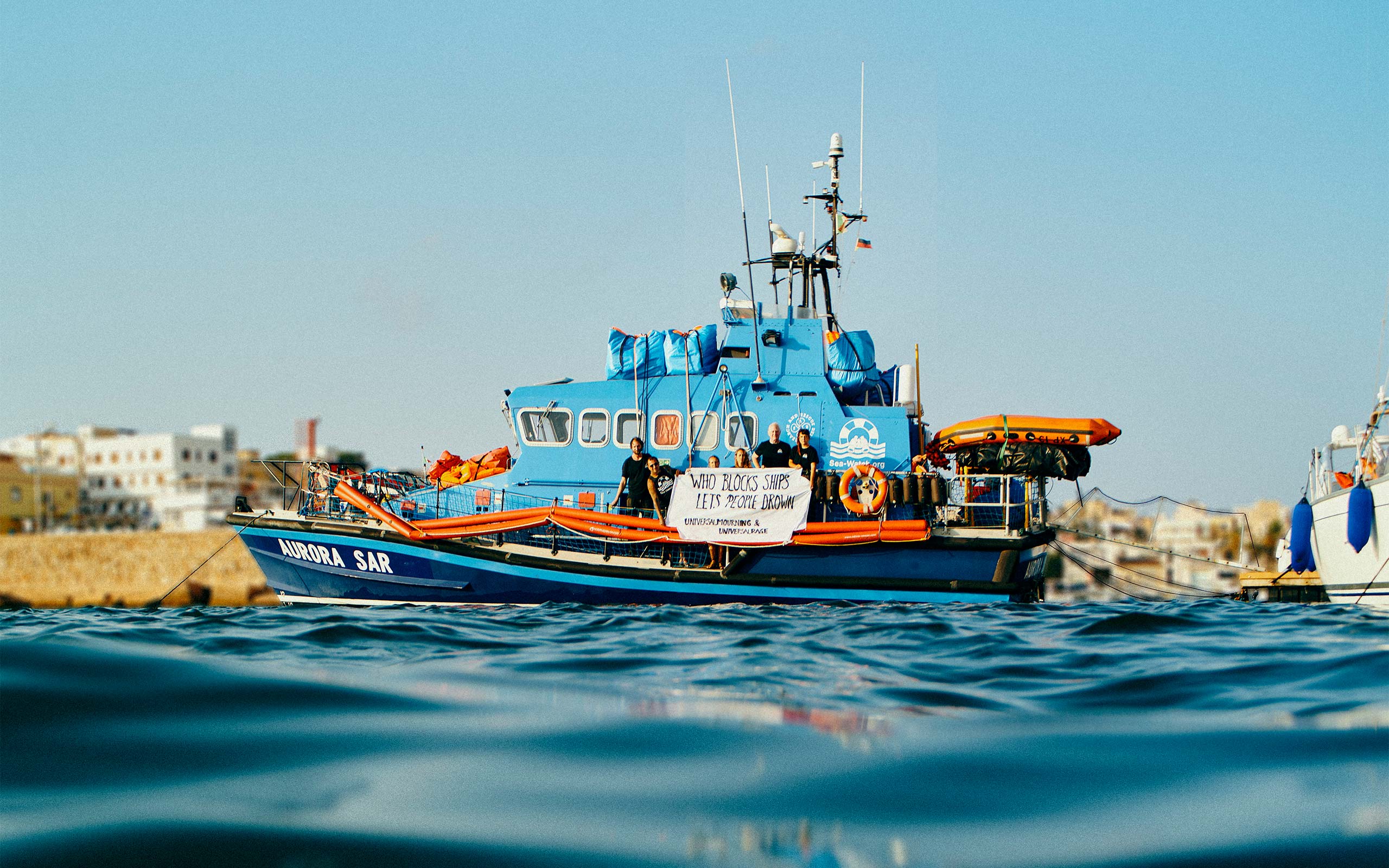 Ein Schiff von Sea-Watch im Mittelmeer. Vorne sind einige Wellen zu sehen, und im Hintergrund einige Häuser, da das Boot sich in Küstennähe befindet.