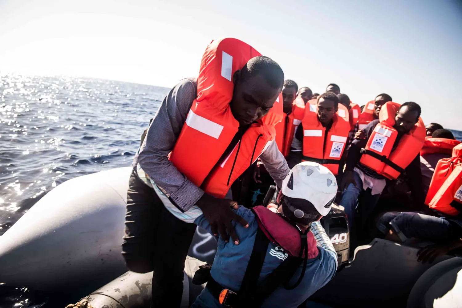 Ein Sea-Watch-Crewmitglied mit Helm hilft einer geretteten Person mit Schwimmweste auf einem überfüllten Schlauchboot im Mittelmeer.