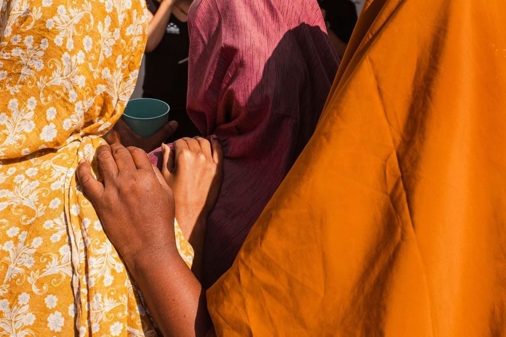 Mehrere Menschen stehen dicht beieinander; man sieht bunte Stoffe in Gelb, Orange und Pink. Eine Hand ruht beruhigend auf der Schulter einer anderen Person – ein Moment der Nähe und Solidarität.