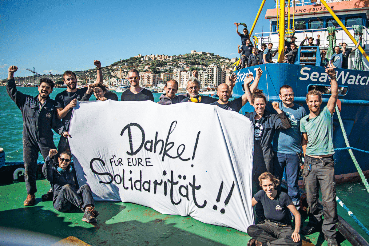 Eine Gruppe von Sea-Watch-Mitgliedern steht auf einem Schiff und hält ein Banner mit der Aufschrift „Danke! Für eure Solidarität!!“. Einige Mitglieder posieren mit erhobenen Fäusten, während andere im Hintergrund winken.