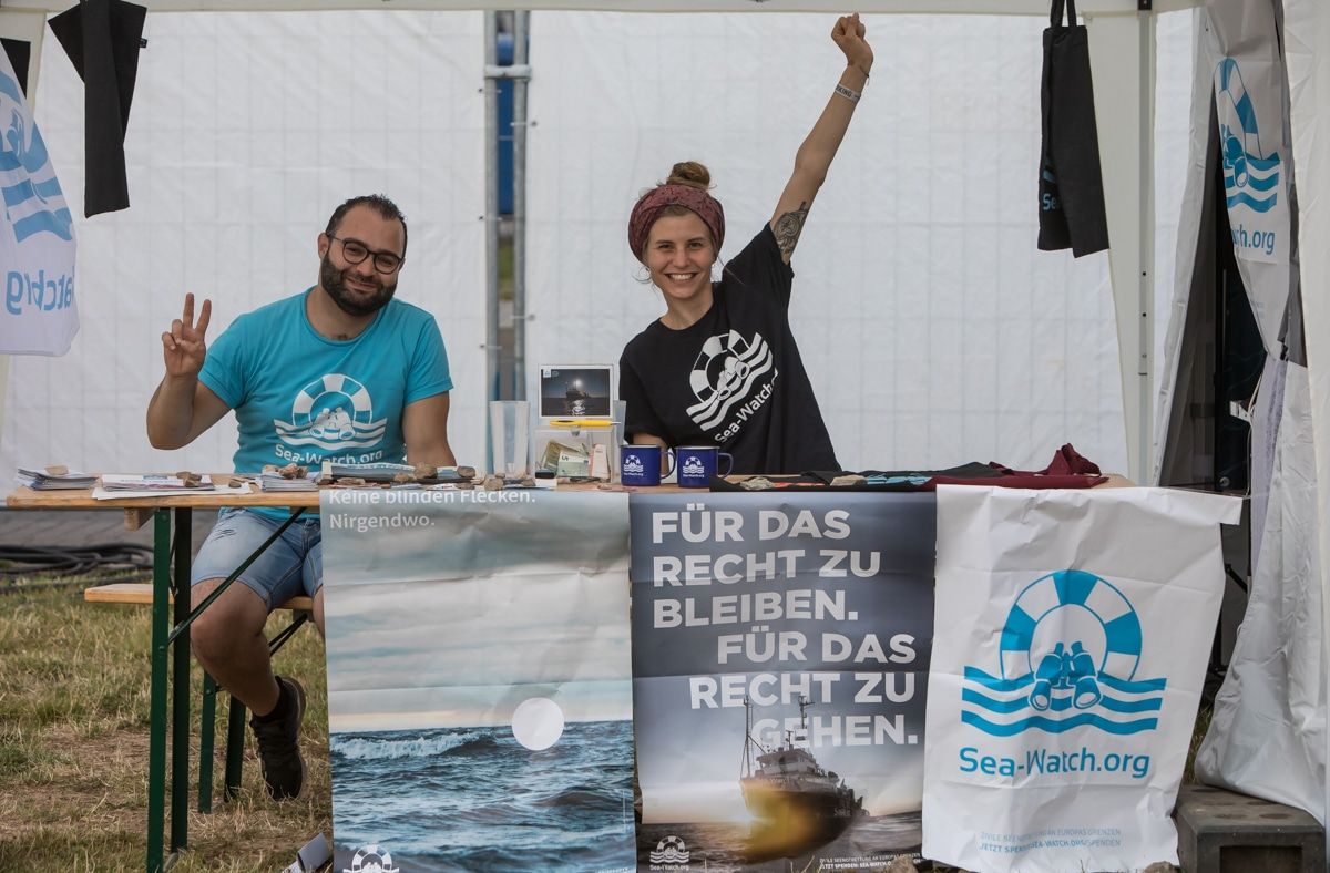 Zwei Personen sitzen an einem Sea-Watch-Stand und posieren motiviert für das Foto. Auf dem Tisch liegen Sea-Watch-Materialien und Plakate.