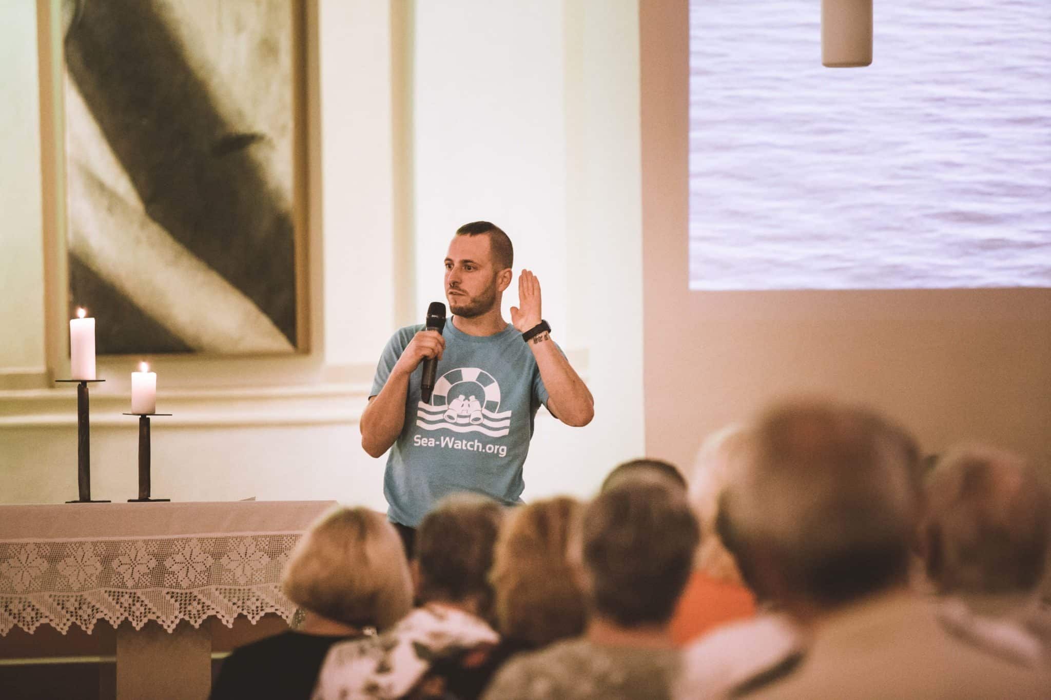 Ein Mann in einem blauen „Sea-Watch.org“-T-Shirt spricht mit einem Mikrofon vor einem Publikum in einem Raum, während im Hintergrund ein Bild von Wasser gezeigt wird. Zwei Kerzen brennen auf dem Tisch vor ihm.