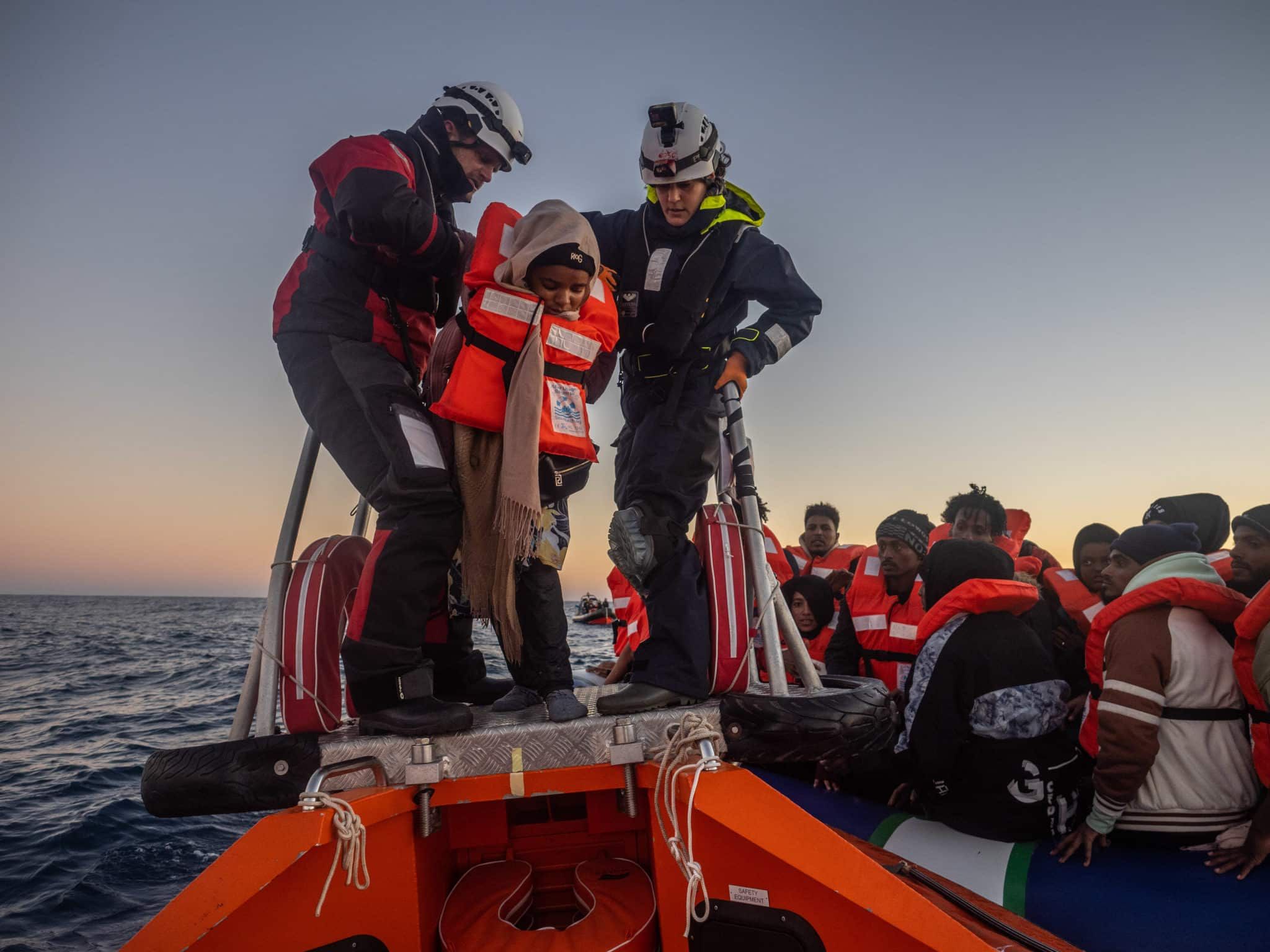 Zwei Seenotretter helfen einer Person in Rettungsweste an Bord eines Rettungsbootes, während eine Gruppe von weiteren geretteten Personen, ebenfalls in Rettungswesten, im Hintergrund sitzt. Das Bild wurde bei Sonnenuntergang auf dem Meer aufgenommen.