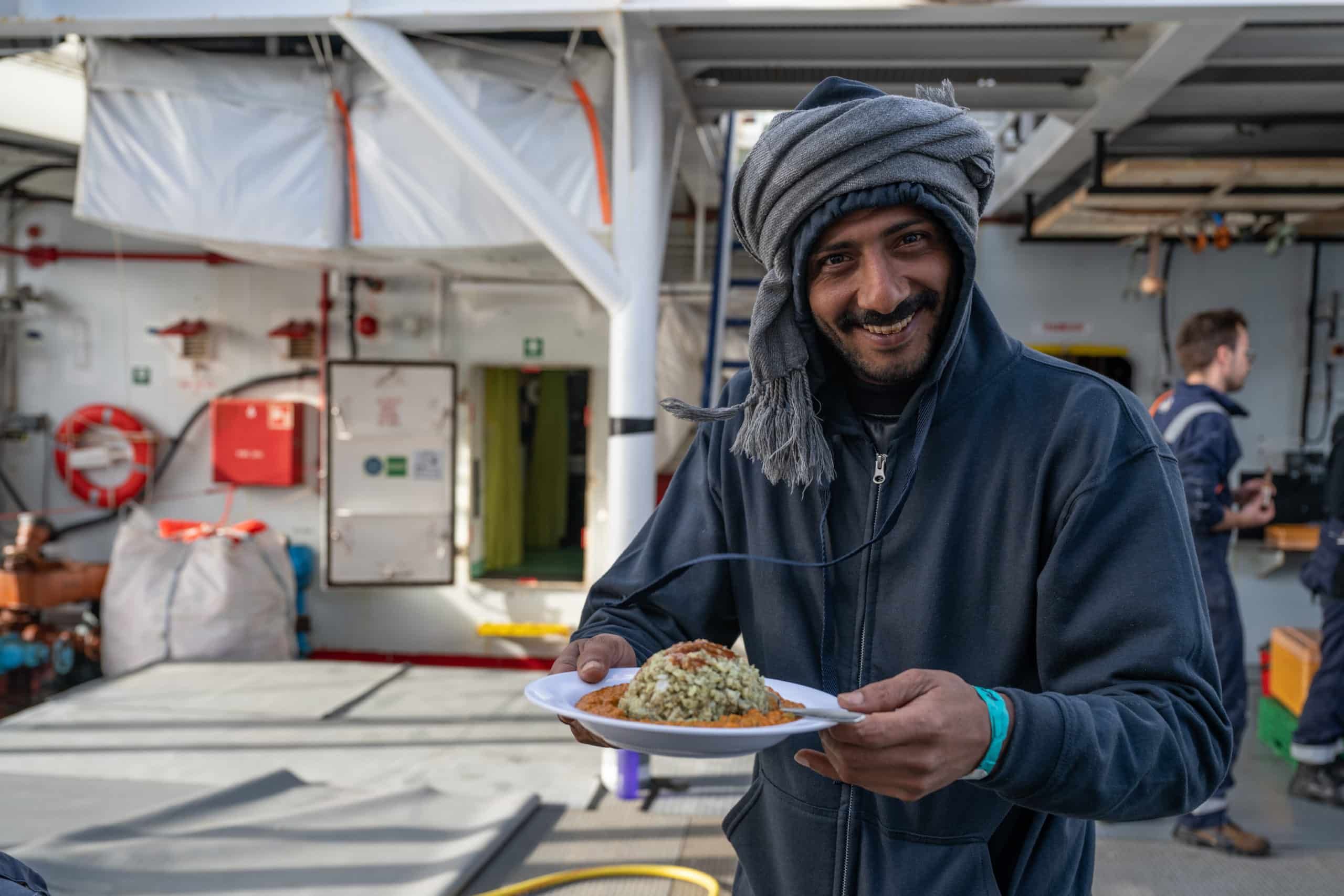 Ein Mann auf einem Schiff hält einen weißen Teller mit Reis und Soße und lächelt in die Kamera. Er trägt einen grauen Hoodie und ein Kopftuch, während im Hintergrund andere Personen zu sehen sind.