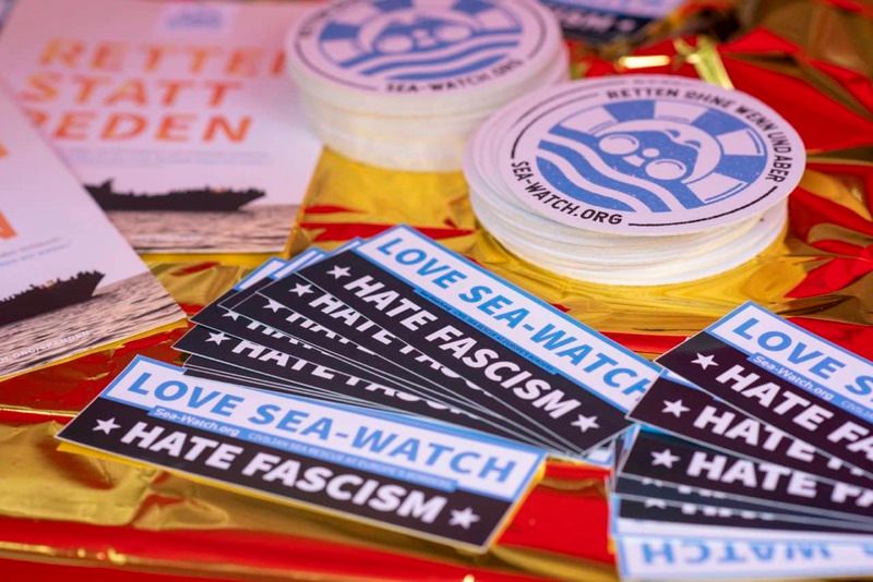 Aufkleber und Flyer von Sea-Watch, erkennbare Slogans „LOVE SEA-WATCH – HATE FASCISM“ und „Retten statt Reden“.