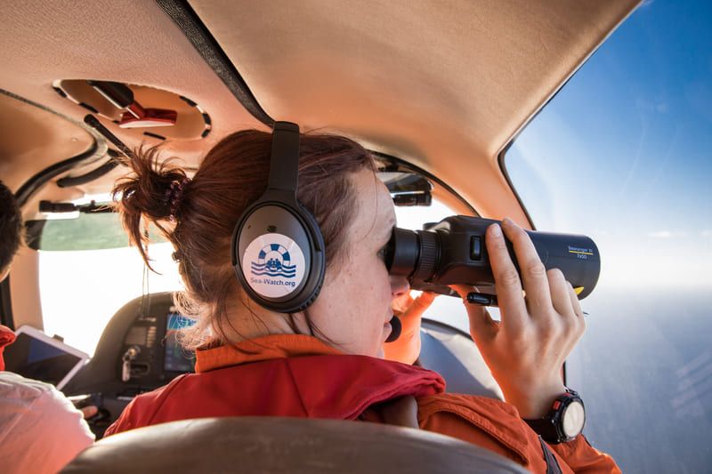 Person mit Headset und Fernglas bei einer Beobachtung aus dem Cockpit eines Flugzeugs, mit Sea-Watch-Logo auf dem Kopfhörer.