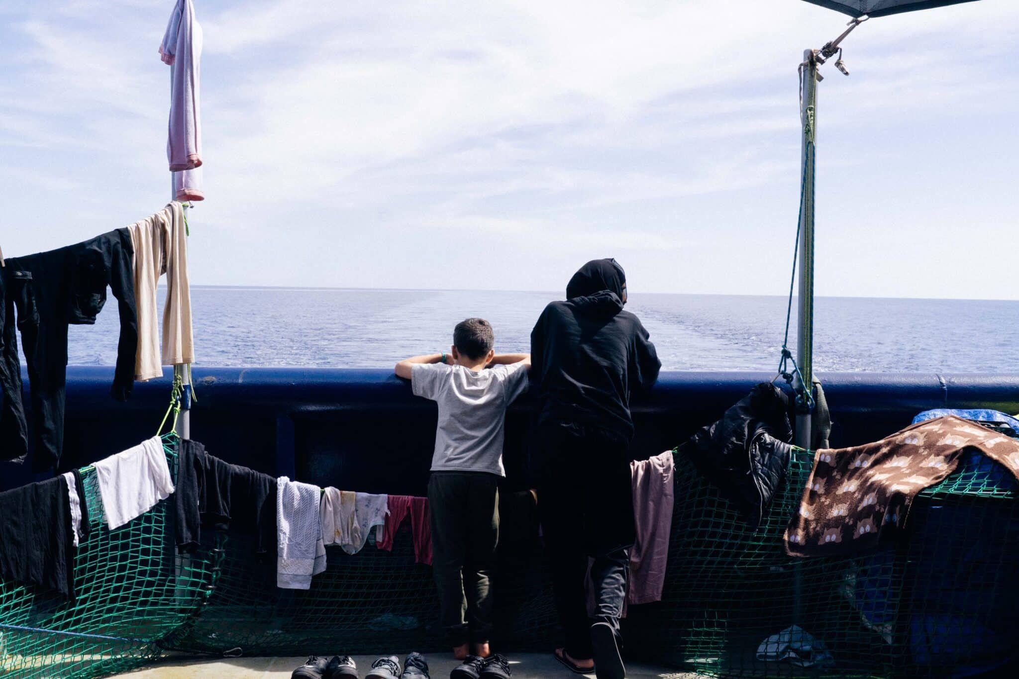 Zwei gerettete Personen stehen an der Reling eines Schiffes und blicken gemeinsam aufs Meer, umgeben von zum Trocknen aufgehängter Wäsche.