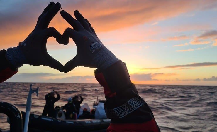 Zwei Hände formen ein Herz vor der Kulisse eines Sonnenuntergangs auf dem Meer – im Hintergrund ein Rettungsboot mit Menschen in Schutzkleidung – ein stilles Zeichen der Solidarität und Hoffnung auf offener See.