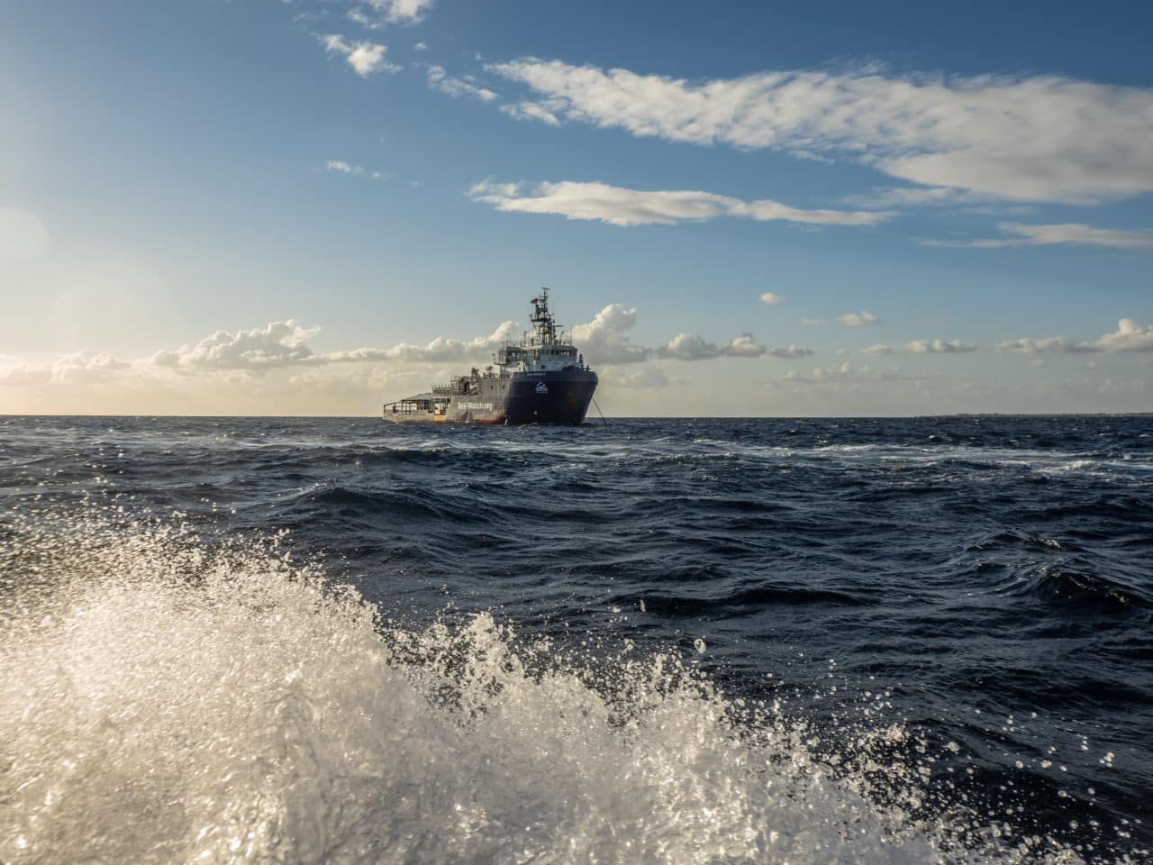Seenotrettungsschiff Sea-Watch 5 auf offenem Meer bei Tageslicht, aufgenommen aus einem kleineren Boot mit spritzender Gischt im Vordergrund.