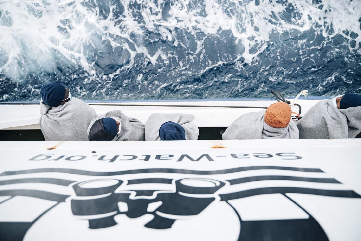Gerettete Menschen in Decken gehüllt sitzen an Bord eines Sea-Watch-Schiffs und blicken auf das Meer.
