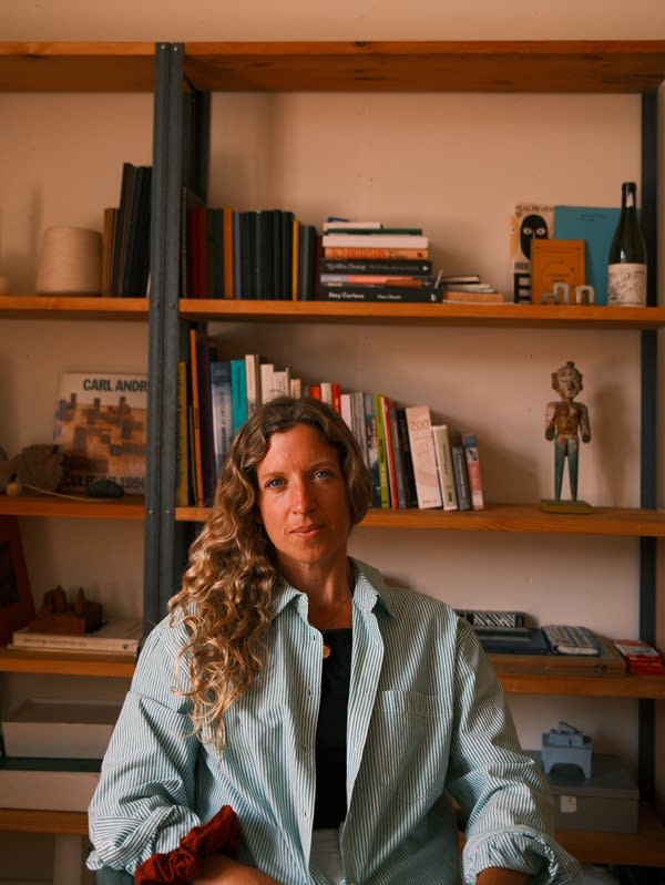 Jess sits in her studio in front of shelves with books and sculptures