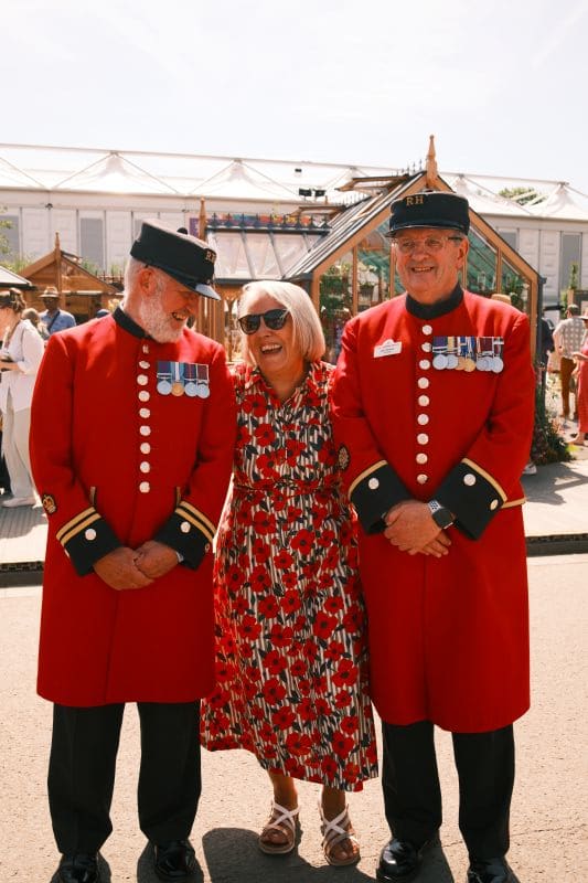 A lady wearing a red Seasalt dress posing with two Chelsea Pensioners in uniform at the Chelsea Flower Show