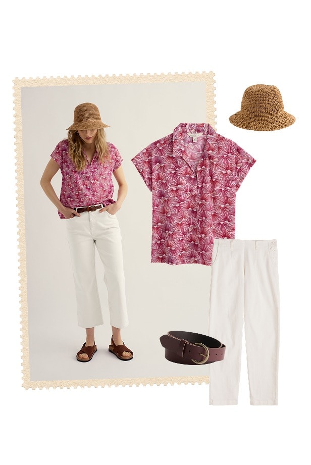 A woman wears a pink shirt with a sunset print. a pair of summery white trousers and a paper sunhat from Seasalt