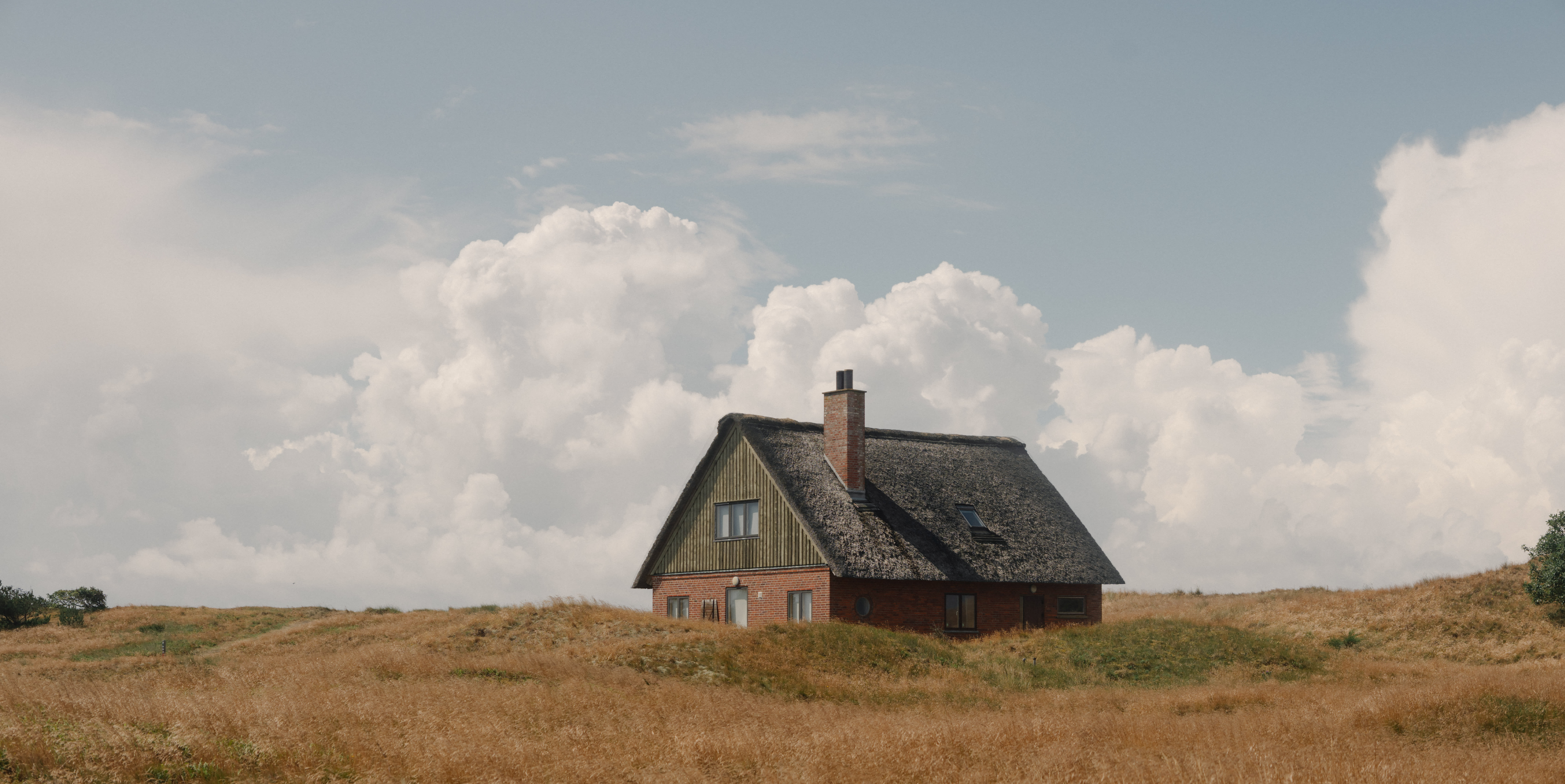 A house stands in a lush field under a dramatic, cloudy sky, creating a serene and picturesque landscape.