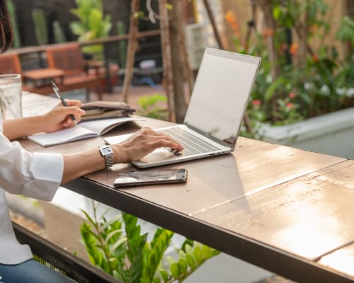 Iemand die buiten op een laptop werkt, aantekeningen maakt in een notitieblok, omringd door groene planten