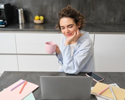 Jeune femme souriante réfléchissant à une idée de business tout en travaillant sur son ordinateur portable, une tasse à la main, dans une cuisine moderne.