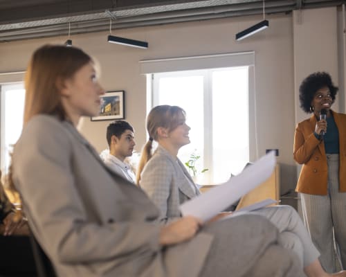 Une femme souriante en veste orange présente avec un micro devant un petit groupe de personnes attentives, lors d’un atelier ou d’une formation en entreprise.