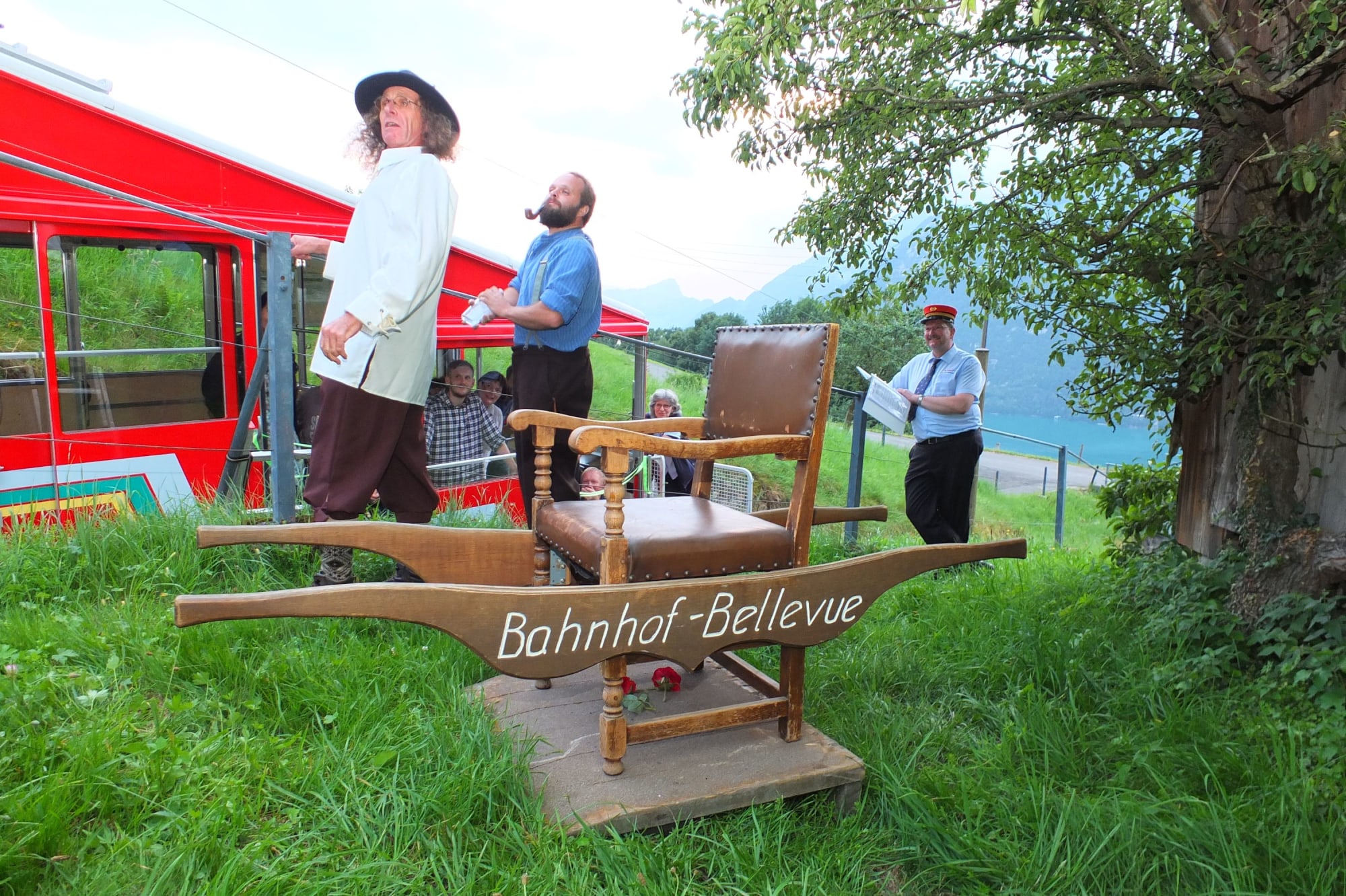 Drei Schauspieler des Bahntheaters auf der Wiese neben dem Bahngleis der Standseilbahn. Im Vordergrund stehet eine Trage  mit einem Sessel für eine Person, angeschrieben mit Bahnhof-Bellevue.