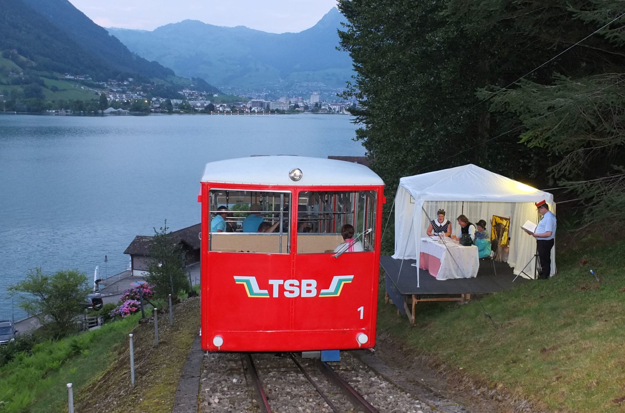 Blick auf einen Wagon der Standseilbahn, im Hintergrund der See und die Berge. Neben dem Gleis ist eine kleine Bühne aufgebaut, gedeckt mit einem seitlich offenen, weissen Pavillon. Vor dem Pavillon steht ein Schauspieler in Bahn-Uniform. Im Pavillon befindet sich ein Tisch mit drei Schauspielenden, zwei Frauen und in der Mitte ein Mann. Sie schauen sich Dokumente an. Hinter ihnen steht ein Abbild des Urner Wappens.