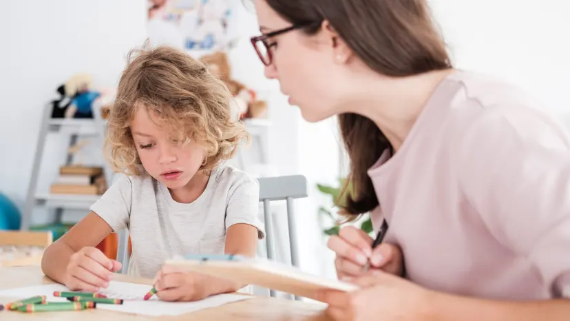 boy with autism sits a play therapy table with his therapist while he colors