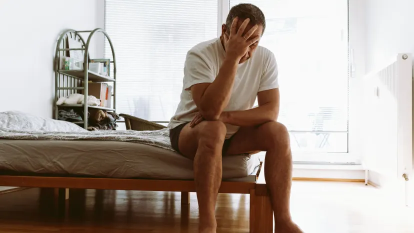Frustrated man suffering from erectile dysfunction sits on his bed with his head in his hand 