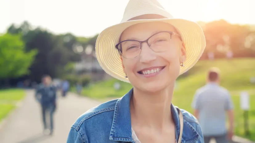 young bald woman who is a cancer survivor wearing a hat and smiling at a park
