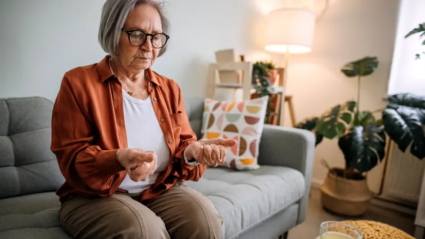 Senior woman sitting on a couch in a living room clutching her fists together due to stiff joints from osteoarthritis