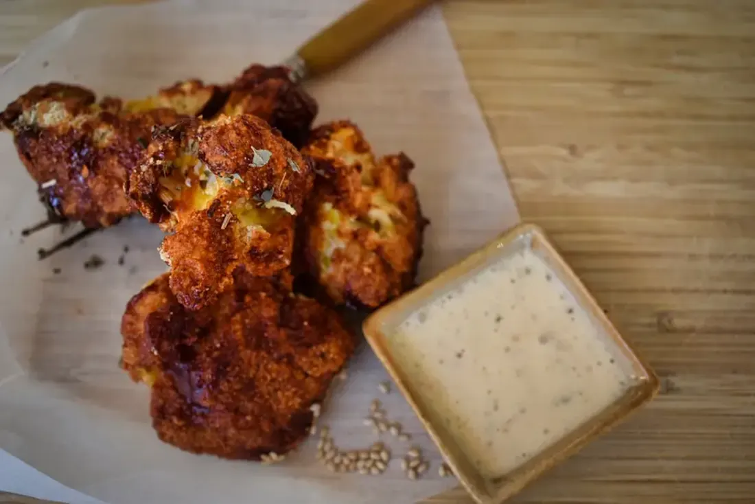 Een houten plank met bakpapier, wings gemaakt van bloemkool met saus erop. Ernaast zie je een bakje saus